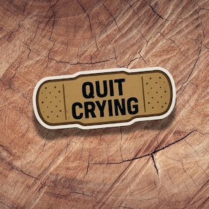 May include: A brown bandage-shaped sticker with the words "QUIT CRYING" in black, outlined in white. The sticker has small dotted details and is set against a textured, brown wooden background.
