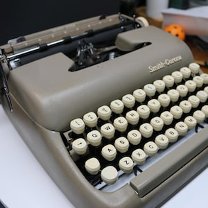 Peut inclure: Une machine à écrire Smith-Corona vintage de couleur grise. La machine à écrire possède un clavier complet avec des touches blanches et des lettres noires. Le nom de la marque est visible à l'avant. La machine est posée sur une surface blanche.