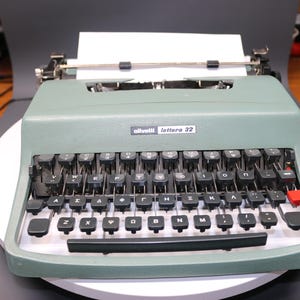 May include: A vintage Olivetti Lettera 32 typewriter in a muted green colour. The typewriter features a full keyboard with black keys, a red return key, and a paper feed mechanism. The model name is visible on the front.