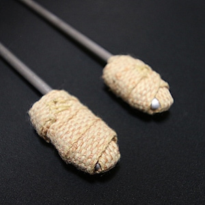 Puede incluir: Dos poi de fuego, accesorios para danza con fuego. Consisten en una varilla metálica gris con una cabeza envuelta de color beige. Las cabezas están hechas de un material tejido, sobre un fondo negro.