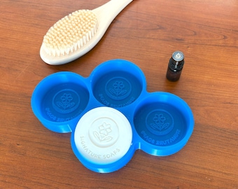 Puede incluir: Un jabonera azul con cuatro compartimentos circulares, uno de ellos con un jabón blanco con el texto "SIGNATURE SOAPS". También hay un cepillo con mango blanco y una pequeña botella marrón. El plato tiene un diseño floral.