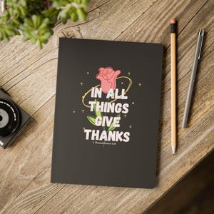 Puede incluir: Un cuaderno negro con el texto "IN ALL THINGS GIVE THANKS" y una ilustración de una rosa rosa. Un lápiz y un bolígrafo están sobre la superficie de madera. El cuaderno está abierto, mostrando la cita de 1 Tesalonicenses 5:18.