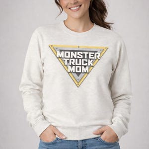 May include: A light gray sweatshirt with the text "MONSTER TRUCK MOM" inside a yellow and silver triangle. The sweatshirt is paired with blue jeans. The woman is smiling.