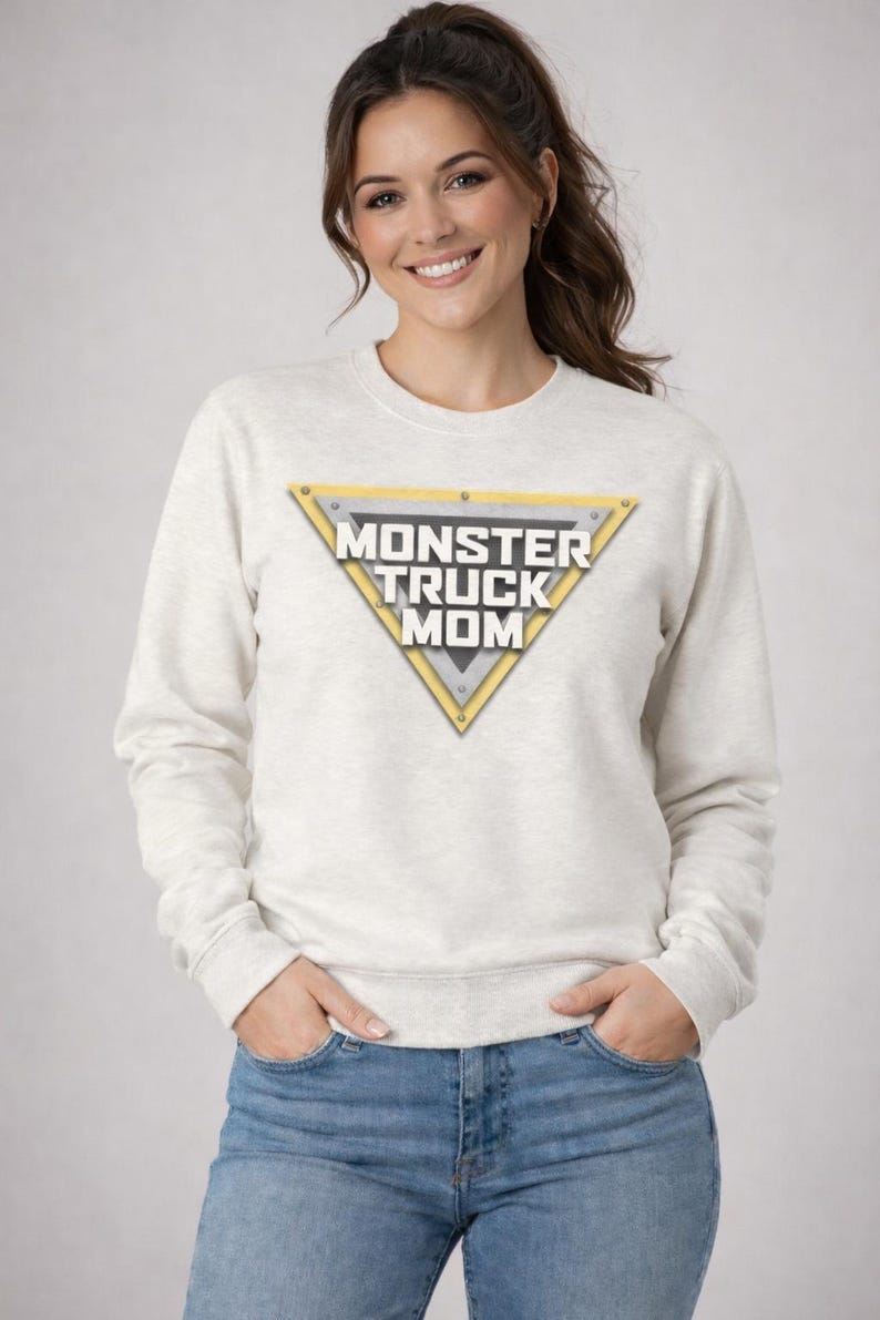 May include: A light gray sweatshirt with the text "MONSTER TRUCK MOM" inside a yellow and silver triangle. The sweatshirt is paired with blue jeans. The woman is smiling.