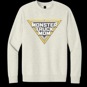 May include: A light beige sweatshirt with a triangular graphic that reads "MONSTER TRUCK MOM" in white and gray. The triangle has a yellow border and is secured with silver rivets. The sweatshirt has long sleeves and a crew neck.