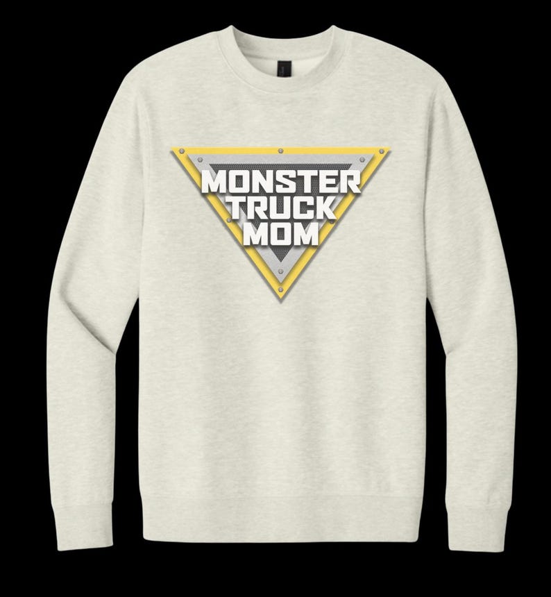 May include: A light beige sweatshirt with a triangular graphic that reads "MONSTER TRUCK MOM" in white and gray. The triangle has a yellow border and is secured with silver rivets. The sweatshirt has long sleeves and a crew neck.