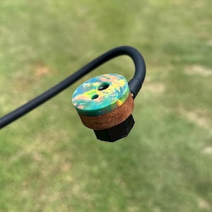 Puede incluir: Un tapón de botella colorido con un mango negro. El tapón presenta un diseño vibrante y arremolinado en tonos verdes, amarillos y azules. Tiene una base de corcho y una parte inferior geométrica negra. El mango es una varilla negra curvada.