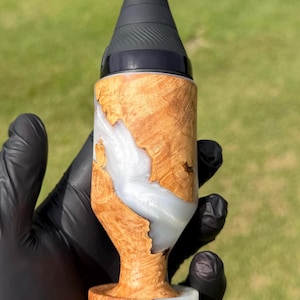 May include: A cylindrical object featuring a combination of natural light brown wood burl and a swirling, pearlescent white resin inlay. The top section is capped with a matte black, tapered component that has a textured, ribbed surface finish.