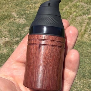 May include: A dark brown, cylindrical wooden object with a black top. The wooden portion has horizontal grooves and a rich, reddish-brown hue. The top is black with a curved design. The object is held in a hand.