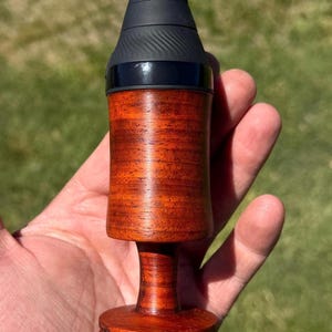 May include: A wooden bottle stopper with a dark finish and a black top. The stopper has a cylindrical body and a flared base. The top is black with a textured grip. The wood grain is visible, adding to its natural aesthetic.