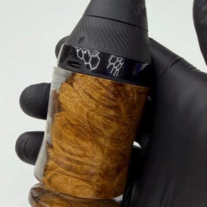 May include: A handheld vaporizer with a black top and a wooden base. The base has a swirling, golden-brown pattern. The vaporizer is held by a gloved hand.