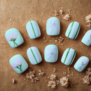 May include: A set of ten artificial nails in a variety of designs. Some nails feature pink flowers, others have pink and green stripes, and two have a bow design. The nails are light blue and are arranged on a brown surface.