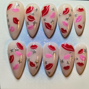 May include: Set of ten almond-shaped false nails. Each nail is painted with a nude base and features red and pink lip designs, along with silver star accents. The nails are arranged in two rows of five.