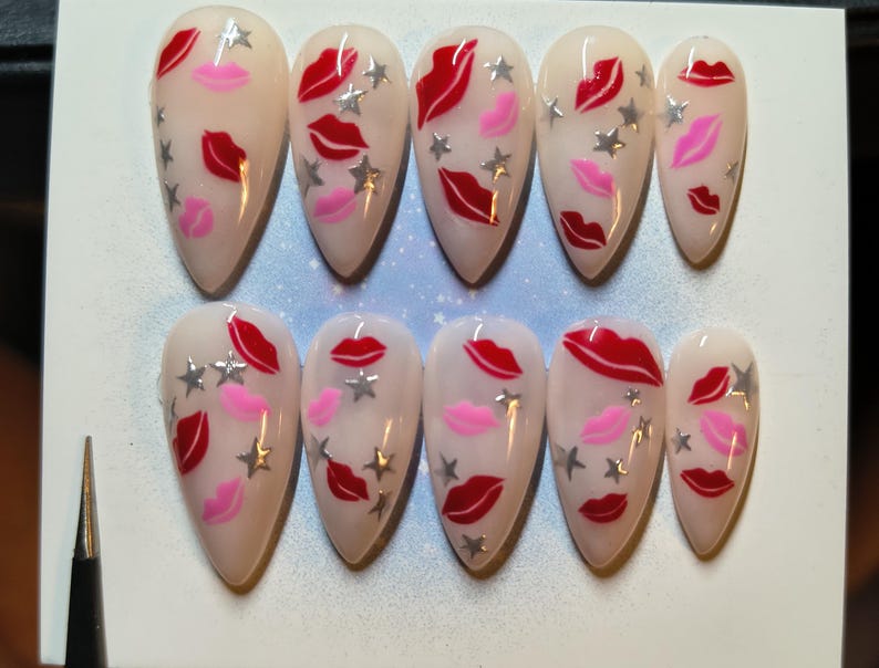 May include: Set of ten almond-shaped false nails. Each nail is painted with a nude base and features red and pink lip designs, along with silver star accents. The nails are arranged in two rows of five.