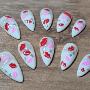 May include: A set of ten white artificial nails, each adorned with a design featuring red lips, pink lips, and silver stars. The nails are arranged on a wooden surface.