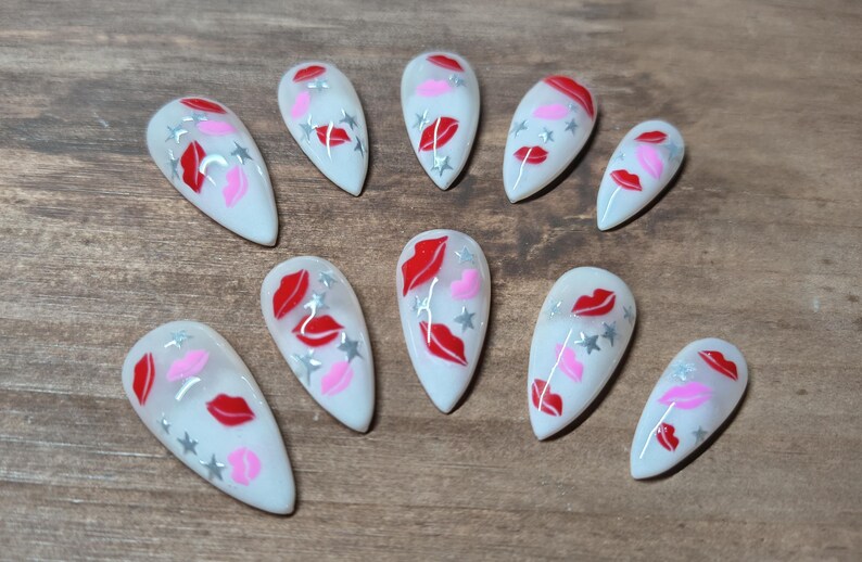 May include: A set of ten white artificial nails, each adorned with a design featuring red lips, pink lips, and silver stars. The nails are arranged on a wooden surface.
