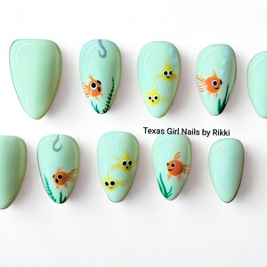 May include: A set of ten almond-shaped press-on nails in a pale mint green base colour. Several nails feature hand-painted orange and yellow goldfish, green seaweed, and small bubbles. The text Texas Girl Nails by Rikki is printed in black.