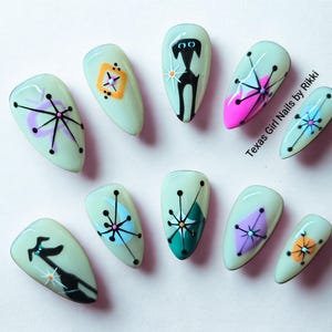 May include: A set of ten almond-shaped press-on nails with a pale mint green base. The designs feature black starbursts, geometric shapes in orange, pink, purple, and teal, and black cat silhouettes. The text Texas Girl Nails by Rikki is visible.