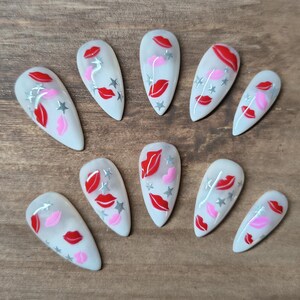 May include: A set of ten white artificial nails, each adorned with a design featuring red lips, pink lips, and silver stars. The nails are arranged on a wooden surface.