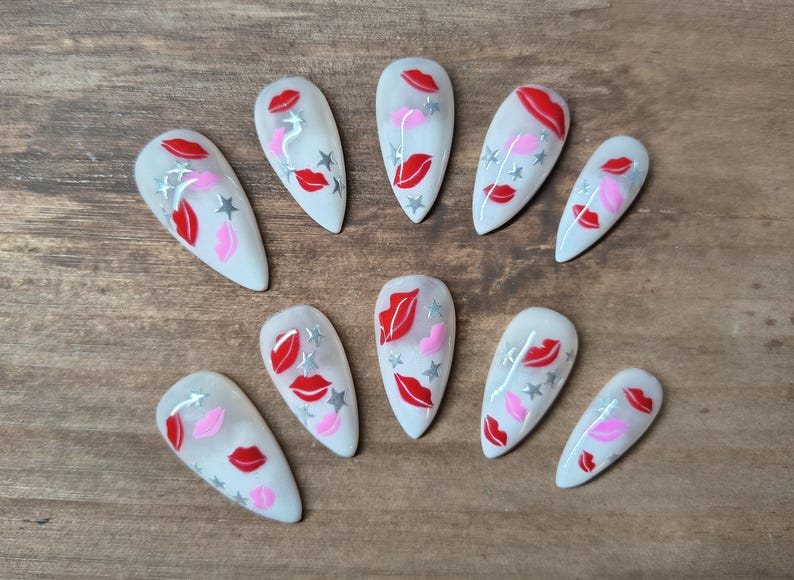 May include: A set of ten white artificial nails, each adorned with a design featuring red lips, pink lips, and silver stars. The nails are arranged on a wooden surface.
