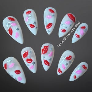 May include: A set of ten white, almond-shaped false nails. Each nail features a design of red and pink lips, silver stars, and the text "Texas Girl Nails by Rikki".
