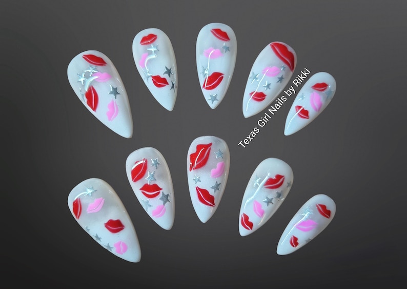 May include: A set of ten white, almond-shaped false nails. Each nail features a design of red and pink lips, silver stars, and the text "Texas Girl Nails by Rikki".