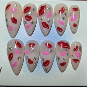 May include: A set of ten almond-shaped false nails. Each nail is painted with a nude base and features red lips, pink lips, and silver star accents. The nails are arranged on a white surface.