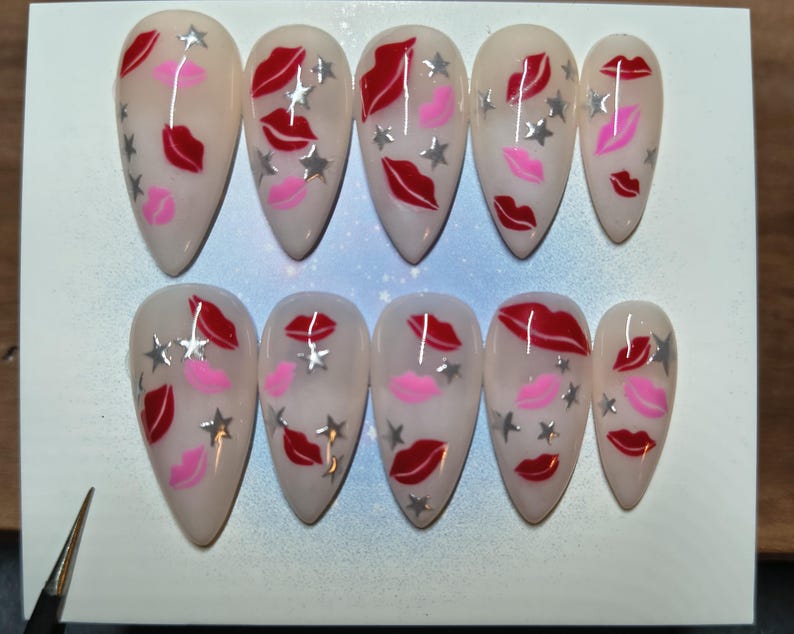May include: A set of ten almond-shaped false nails. Each nail is painted with a nude base and features red lips, pink lips, and silver star accents. The nails are arranged on a white surface.