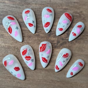 May include: A set of ten white, almond-shaped artificial nails. Each nail features a design of red and pink lips, silver stars, and silver accents. The nails are arranged on a wooden surface.