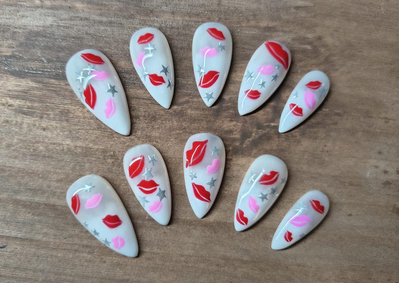 May include: A set of ten white, almond-shaped artificial nails. Each nail features a design of red and pink lips, silver stars, and silver accents. The nails are arranged on a wooden surface.