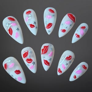 May include: A set of ten white, almond-shaped artificial nails. Each nail is decorated with red and pink lips, silver stars, and a few pink hearts. The nails are arranged on a dark gray background.