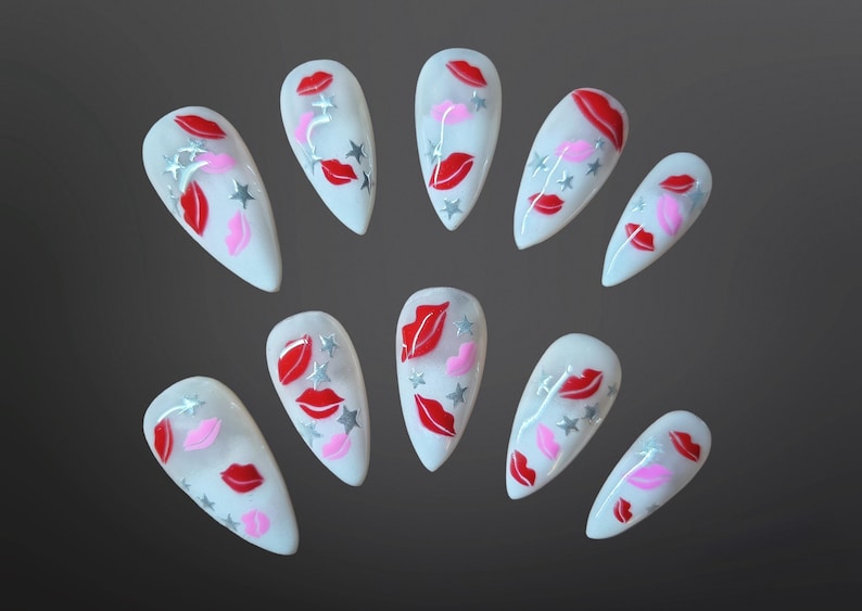 May include: A set of ten white, almond-shaped artificial nails. Each nail is decorated with red and pink lips, silver stars, and a few pink hearts. The nails are arranged on a dark gray background.