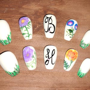 May include: A collection of ten white, oval-shaped artificial nails. Some nails feature floral designs in purple, orange, and blue. Others display the letters "B" and "H" in black script. Green grass-like accents are also present.