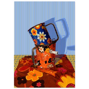 Op de afbeelding: Twee vintage emaille bekers met bloemenpatronen, gestapeld op elkaar. De bovenste beker is blauw met witte en oranje bloemen. De onderste beker is oranje met zwarte en witte bloemen. De bekers staan op een bruin, gepatroneerd tafelkleed.