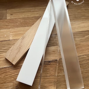 Könnte beinhalten: Drei rechteckige Materialstücke: ein hellbraunes Holzstück, ein weißes Stück und ein klares Acrylstück. Das Acrylstück ist das größte und leicht angewinkelt. Das Holzstück ist das kleinste und liegt flach. Das weiße Stück liegt dazwischen und liegt ebenfalls flach. Das Acrylstück hat einen leichten Glanz.