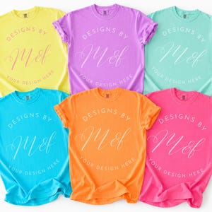 May include: Six short-sleeved crew neck t-shirts in yellow, purple, teal, blue, orange, and pink. Each shirt features a circular graphic with the text 'DESIGNS BY M&f' and 'YOUR DESIGN HERE' printed in a contrasting color on the center of the chest.