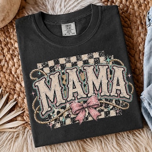Puede incluir: Camiseta negra con la palabra "MAMA" en una fuente de estilo occidental, un lazo rosa y detalles de cuerda. El diseño incluye un patrón de cuadros y estrellas. La camiseta se muestra sobre una superficie tejida.