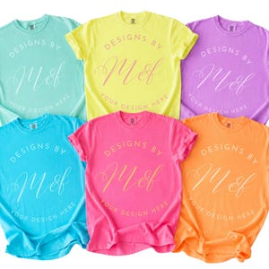 May include: A collection of five short-sleeved crew neck t-shirts in mint green, yellow, lavender, bright blue, hot pink, and orange. Each shirt features a circular graphic with the text "DESIGNS BY M&f YOUR DESIGN HERE" printed in white.