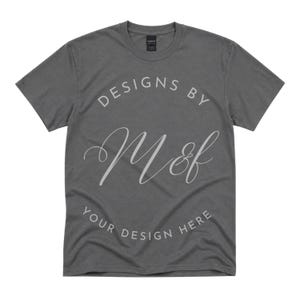 Może przedstawiać: Ciemnoszary t-shirt z krótkim rękawem z napisem "DESIGNS BY" w łukowatym układzie nad dużym logo "M&f" pisanym kursywą, z napisem "YOUR DESIGN HERE" wydrukowanym w zakrzywionym formacie wzdłuż dolnej części projektu graficznego.
