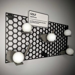 May include: A black metal honeycomb wall organizer with golf balls and golf clubs. The organizer has a sign that reads "GOLF [gawif] noun An endless series of tragedies obscured by the occasional miracle."