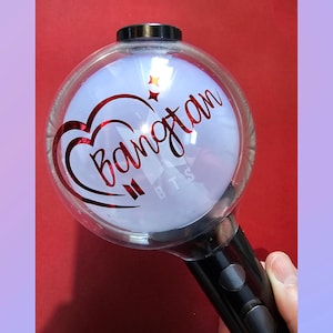 May include: A spherical light stick with a clear outer shell and a white inner globe. The surface features red metallic text reading Bangtan, a red heart outline, two small red stars, and the white letters BTS at the bottom. A black handle is attached.