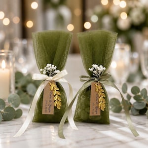 May include: Two olive green tulle favor bags tied with cream ribbons, each adorned with a sprig of white baby's breath, a gold-toned leaf charm, and a rectangular kraft paper tag printed with the text "Cinini & Giegin 31.05.2025".