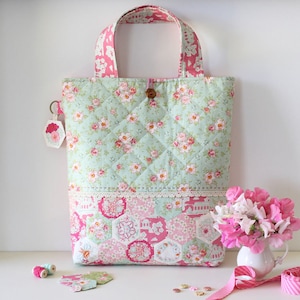Könnte beinhalten: Eine gesteppte Tragetasche mit einem Blumendruck in Rosa- und Grüntönen. Die Tasche hat einen braunen Knopfverschluss und einen pink-weiß gemusterten Trageriemen. Die Tasche steht auf einer weißen Oberfläche mit einem kleinen Strauß rosa Blumen in einer weißen Vase.