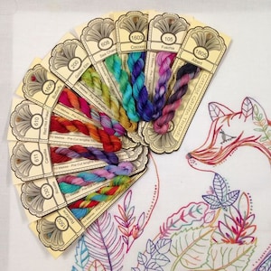 May include: A fan of 10 skeins of embroidery floss in various colours, including red, orange, yellow, green, blue, purple, and pink. Each skein is labelled with a number and a colour name. The skeins are arranged in a fan shape, with the labels facing outwards. The skeins are all 10 metres long and pre-cut. The text on the labels reads "Cottage Garden Threads".