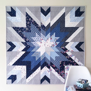 Op de afbeelding: Een blauwe en witte quilt met een stervormig patroon. De quilt is gemaakt van verschillende tinten blauw en witte stoffen, en het stervormige patroon is gemaakt met verschillende geometrische vormen. De quilt hangt aan een witte muur.