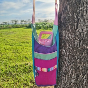 Peut inclure: Un sac fourre-tout en tissu coloré avec une bouteille d'eau rose à l'intérieur. Le sac a un design bleu sarcelle, violet, vert et rose avec une poche zippée sur le devant. Le sac est suspendu à un arbre.