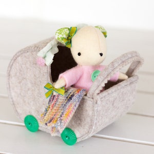 May include: A handmade doll in a gray felt stroller. The doll wears a pink shirt and has green and yellow bows in its hair. A small, colorful knitted blanket rests in the stroller. The stroller has green wheels.