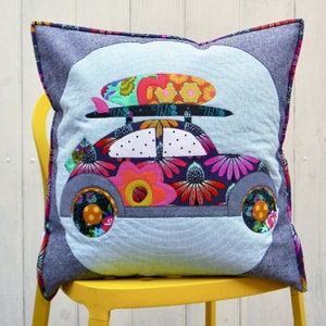 Puede incluir: Cojín decorativo cuadrado con un diseño de coche caprichoso con una tabla de surf encima. El coche está adornado con coloridos estampados florales y un fondo azul claro. El cojín está enmarcado con una tela similar al denim.