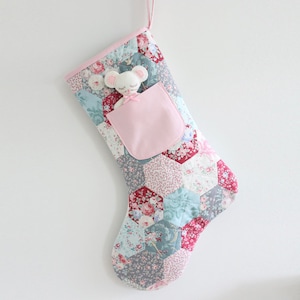 May include: A pink and blue patchwork Christmas stocking with a pink pocket and a white stuffed mouse inside.