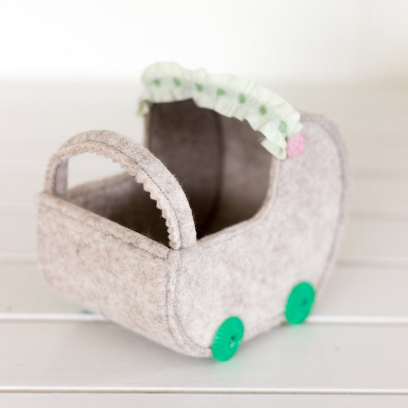 Peut inclure: Une poussette pour poup&eacute;e en feutre gris clair avec une poign&eacute;e festonn&eacute;e et des roues vertes en forme de bouton. La poussette a un auvent vert clair et rose &agrave; volants. Un jouet charmant pour les enfants.